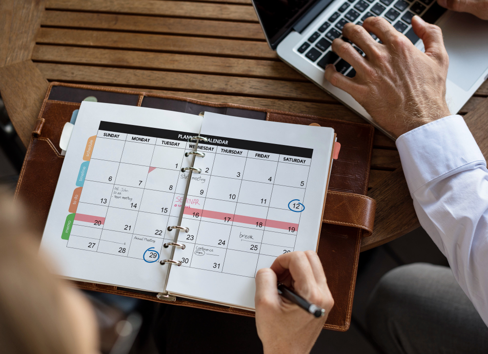 Agenda con calendario che viene compilato a penna. Sullo sfondo mani maschili digitano su un portatile